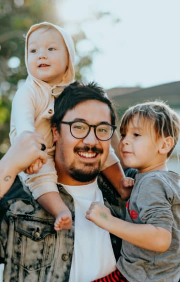 A photo of a dad with his two young children, one sitting on his shoulders and the other on his hip.